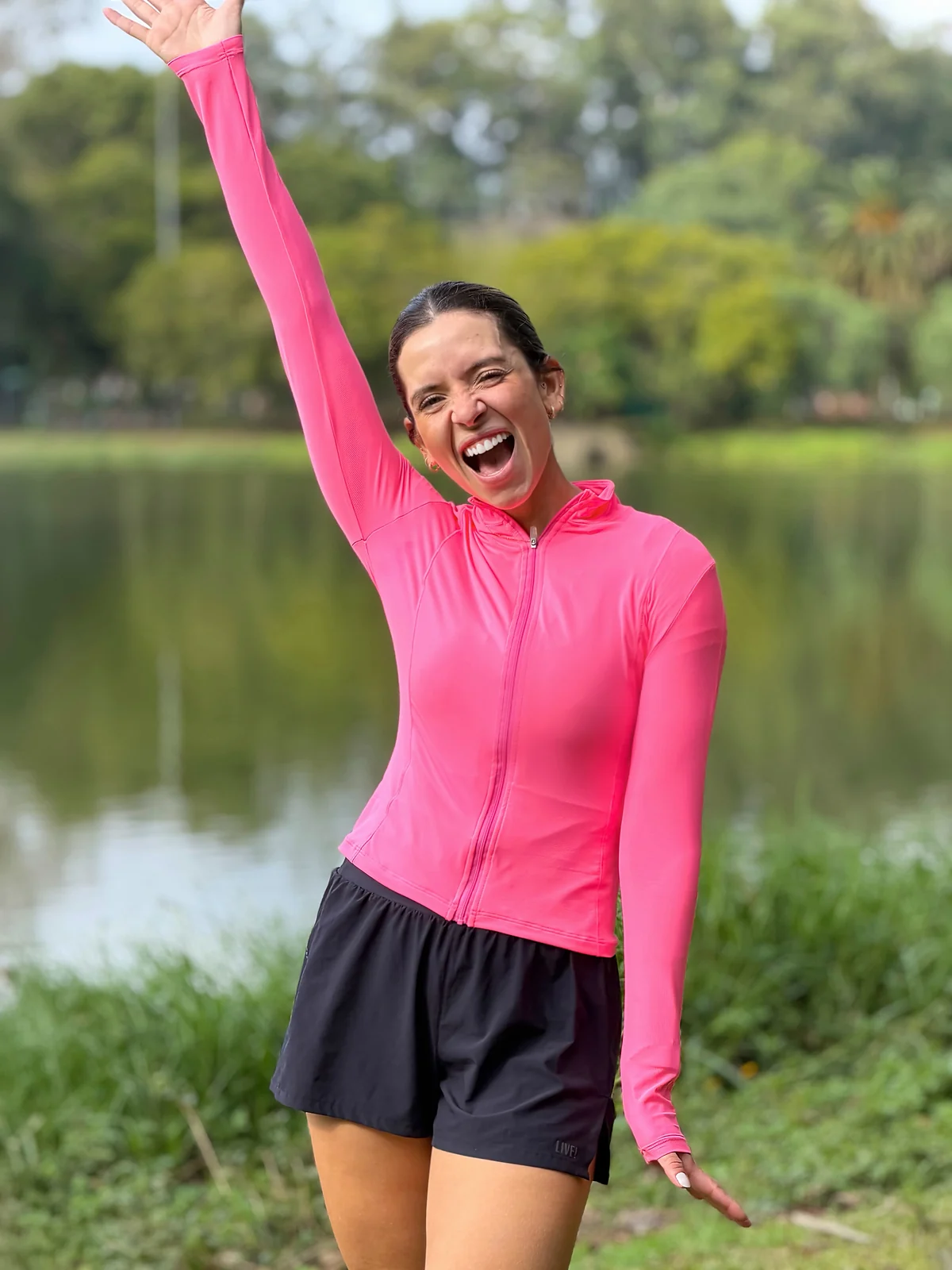 Sthéfany rindo com um braço para cima, de jaqueta rosa de corrida, em frente a um lago