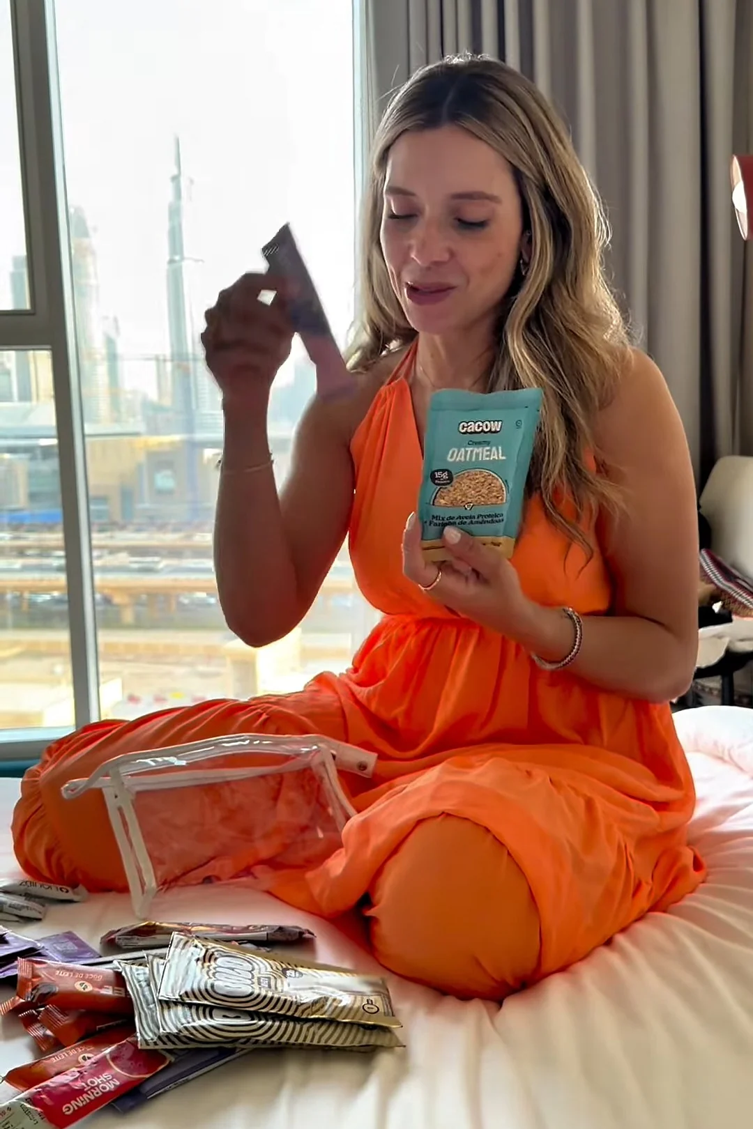 Sthéfany de vestido laranja sentada na cama, mostrando um pacote de aveia da sua necessaire de suplementos