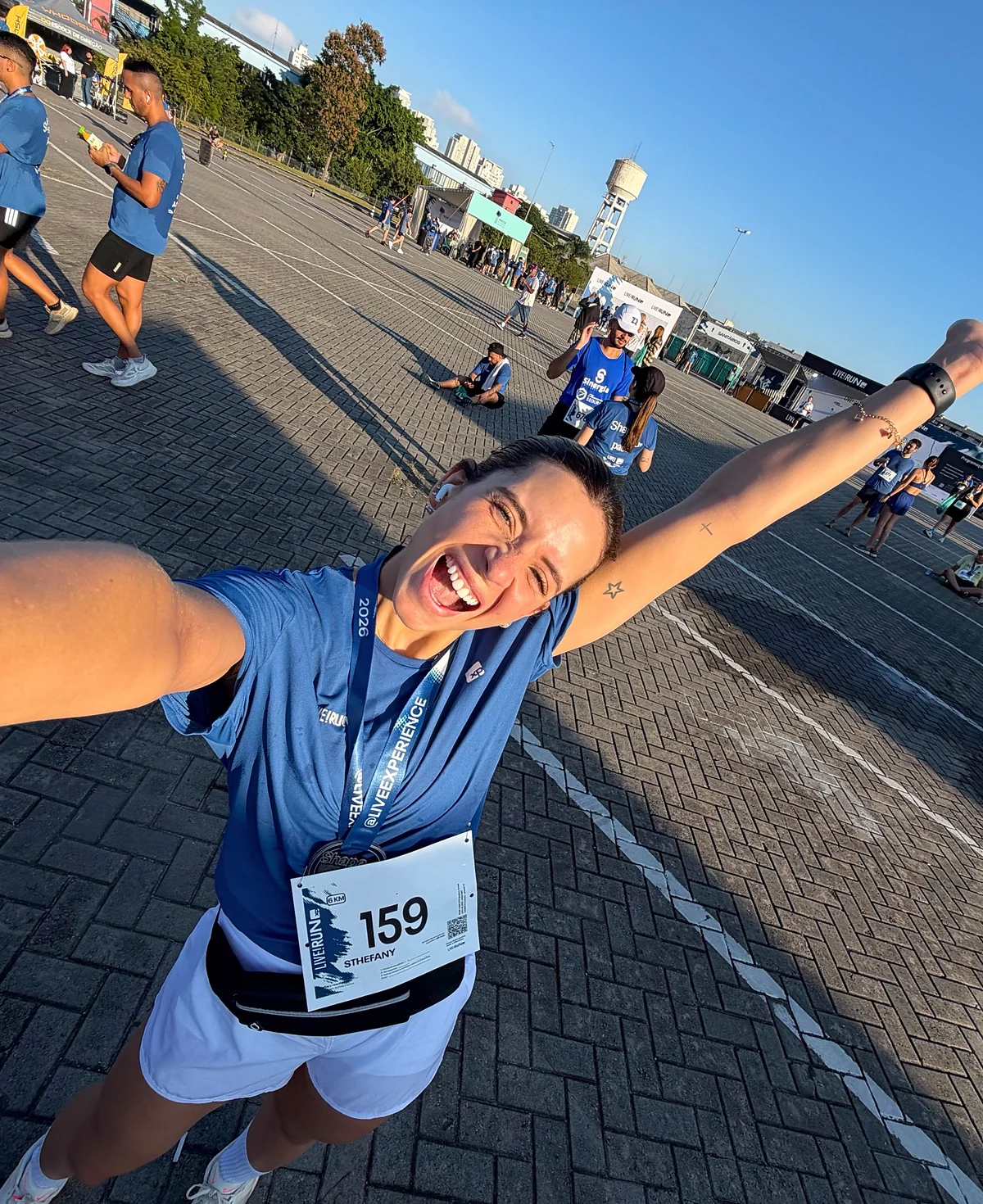 Sthéfany de braços abertos, sorrindo, com camisa azul e número de prova 159, em uma avenida fechada para corrida