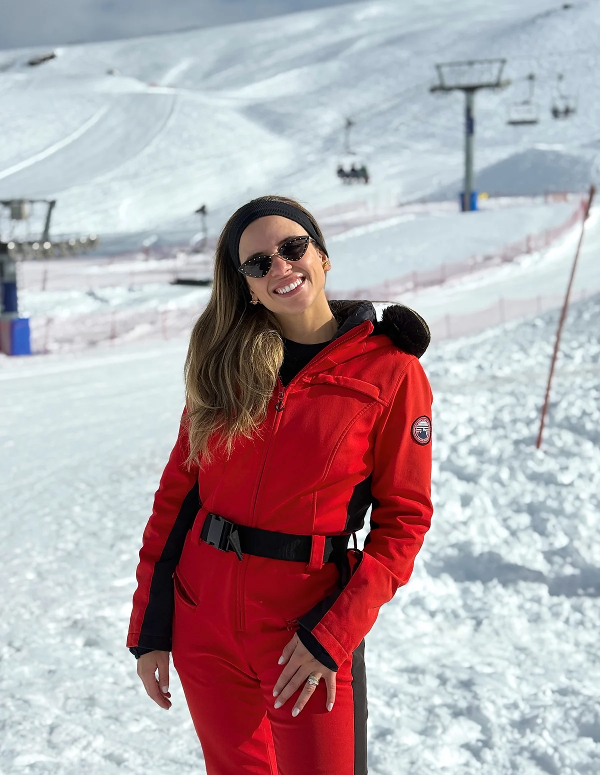 Sthéfany de macacão de esqui vermelho e óculos escuros, na neve, com pista e teleférico ao fundo